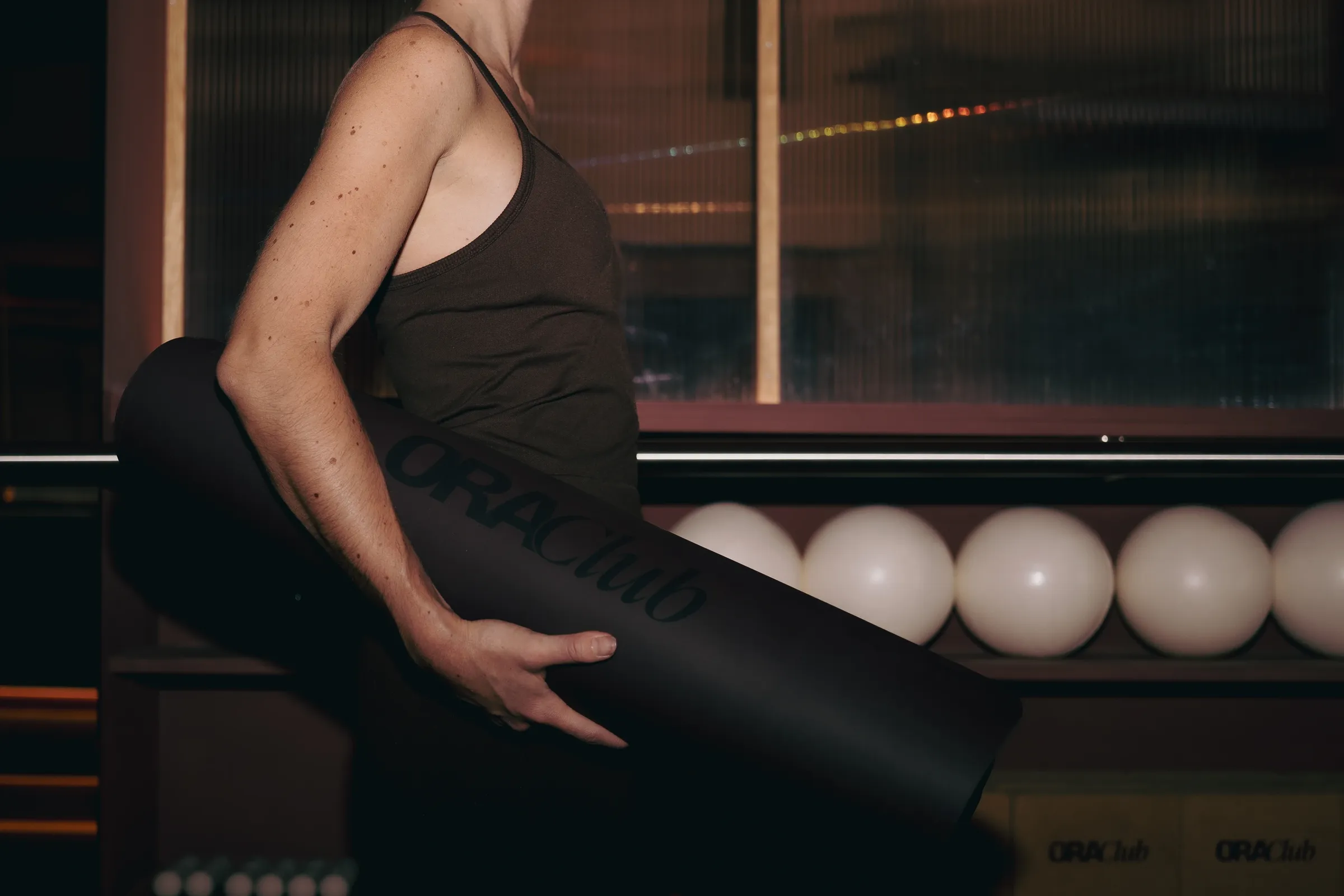 Pratiquante portant un tapis Ora Club roulé devant les étagères du studio